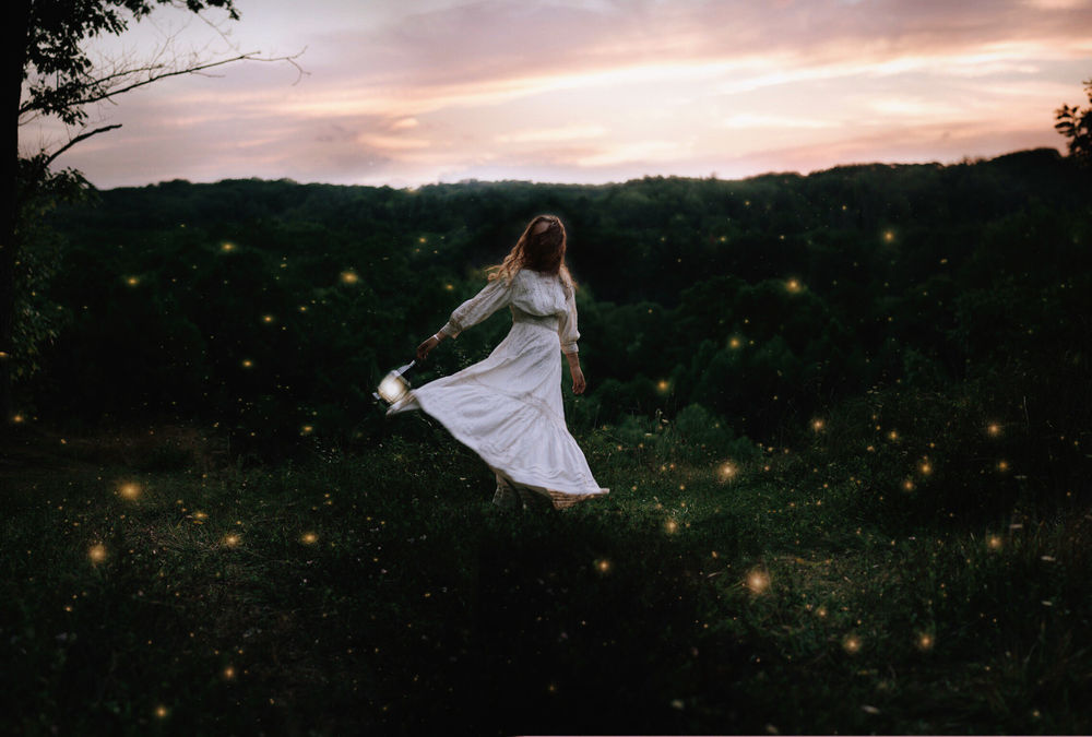 Обои для рабочего стола Девушка, одетая в белое платье, с фонарем в руках, танцует на зеленой поляне в окружении светлячков