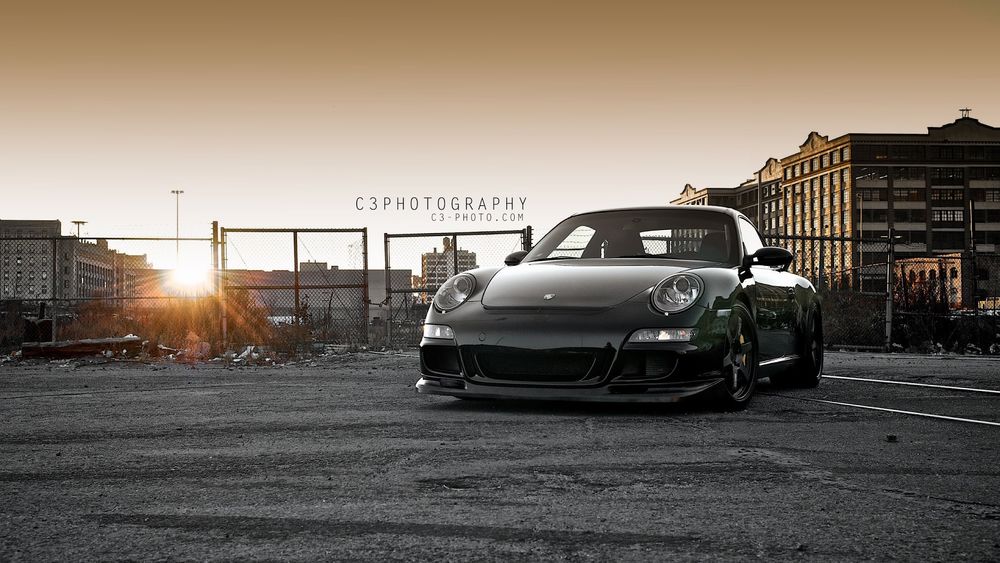 Обои для рабочего стола Черная Porshe 911 на фоне домов и неба