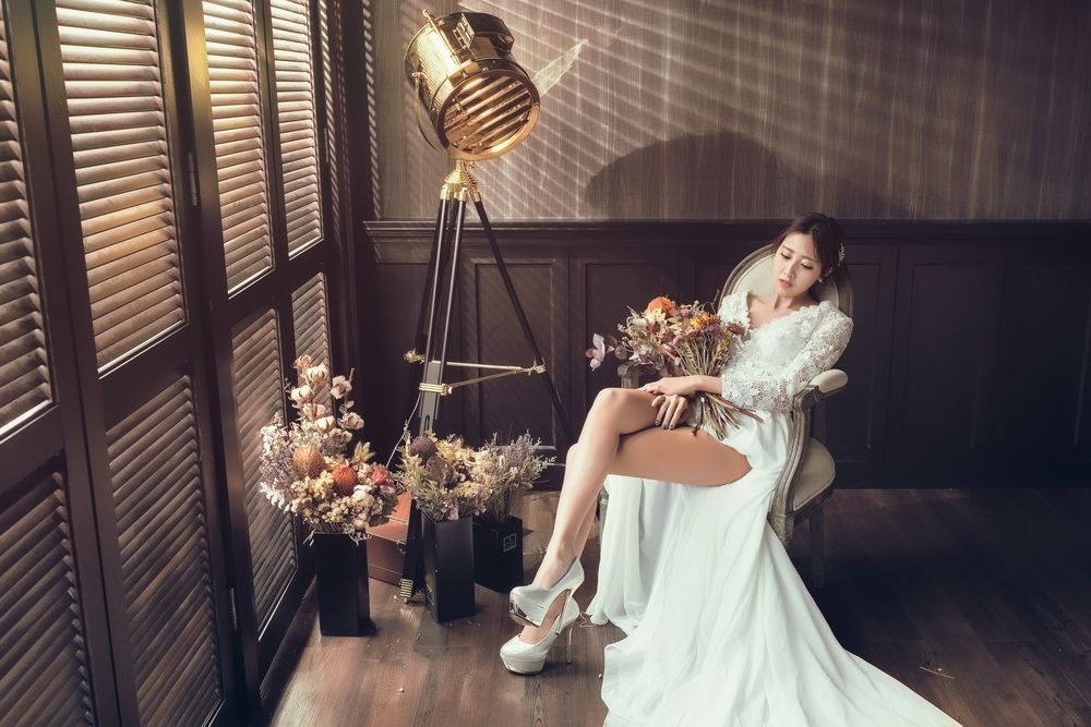 Обои для рабочего стола Азиатка в свадебном платье с букетом сидит на стуле рядом цветы в вазах и софит на треноге