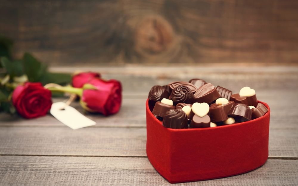 Обои для рабочего стола Шоколадные конфеты в красной коробке в виде сердца, рядом лежат алые розы