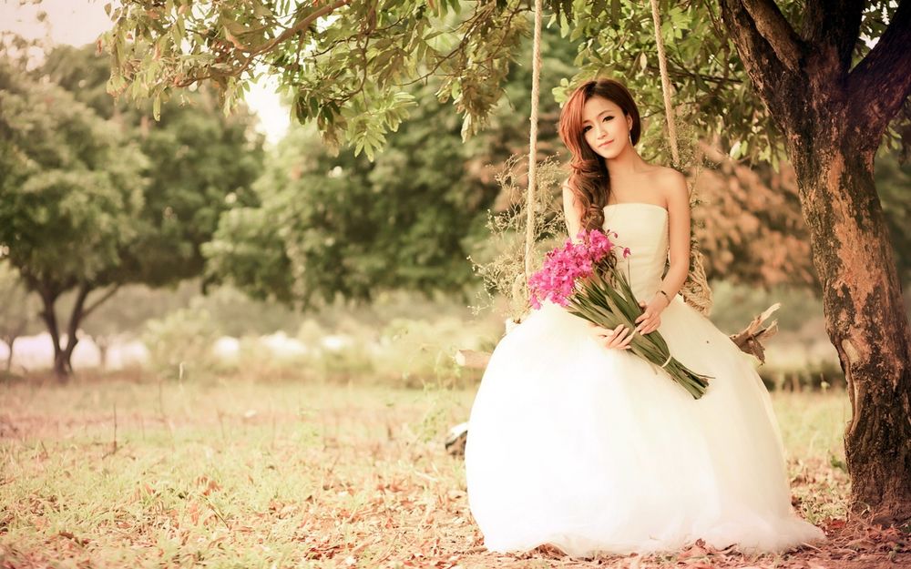 Обои для рабочего стола Девушка азиатка в свадебном платье с букетом цветов в руках сидит на качели