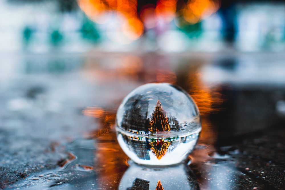 Обои для рабочего стола В стекляном шаре, стоящем в луже на земле, отражается городская улица с новогодней елкой