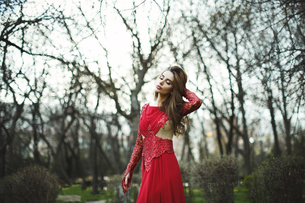 Обои для рабочего стола Девушка красивая шатенка в красном полу ажурном платье стоит на фоне весеннего пейзажа опустив глаза и поправляя свои волосы, by Tamerlan Kagermanov