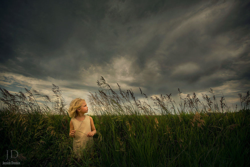 Обои для рабочего стола Девочка стоит на лугу, смотря встревоженно на облачное небо, держа травинку в руке, by Djessica Drossin