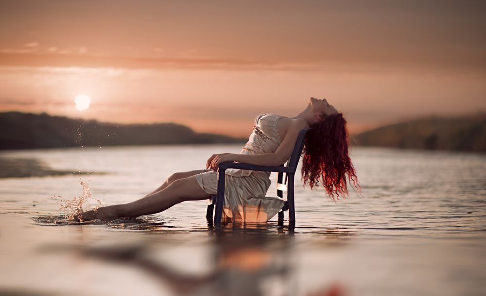 Обои для рабочего стола Рыжеволосая девушка сидит, наслаждаясь природой, погрузив красивые ноги в ласковые волны моря и любуясь заходящим солнцем, медленно уходящем за горизонт