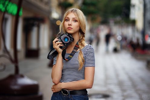 Обои на рабочий стол Модель Алиса Тарасенко / Alice Tarasenko с косичками в полосатом топе и шортах с фотоаппаратом в руке задумавшись стоит на размытом фоне города. Фотограф Ден Евдокимов / DBond
