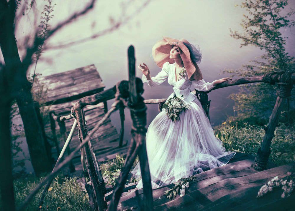 Обои для рабочего стола Красивая девушка в белом длинном платье цветами, в шляпе стоит на ступенях деревянной лестницы, ведущей на мост к воде, фотограф Валерия Мытник