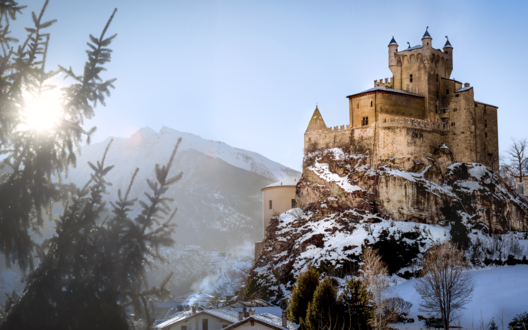 Обои на рабочий стол Заснеженный старинный Castello di Saint-Pierre, Italy / замок Сен-Пьер, Италия на скале на фоне пробивающегося солнца и размытых очертаний гор