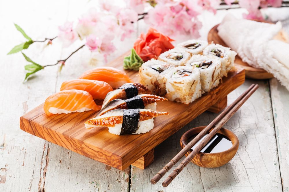 Обои для рабочего стола Японская кухня: суши и роллы, маринованный имбирь и васаби на деревянной подставке, рядом полочки для еды и ветка сакуры