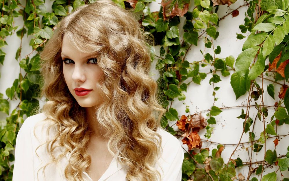 Обои для рабочего стола Певица Тейлор Свифт / Taylor Swift с длинными волнистыми волосами, стоит на фоне стены, увитой плющом