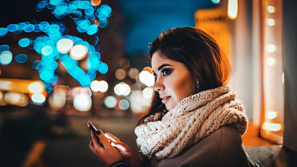 Обои для рабочего стола Красивая девушка в руке с мобильным телефоном, смотрит в сторону, на фоне ночных огней города. Фотограф Антон Харисов