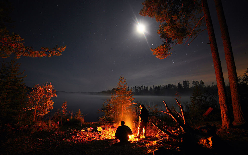 Обои для рабочего стола Двое мужчин возле горящего костра на фоне ночного неба, озера и освещенных деревьев