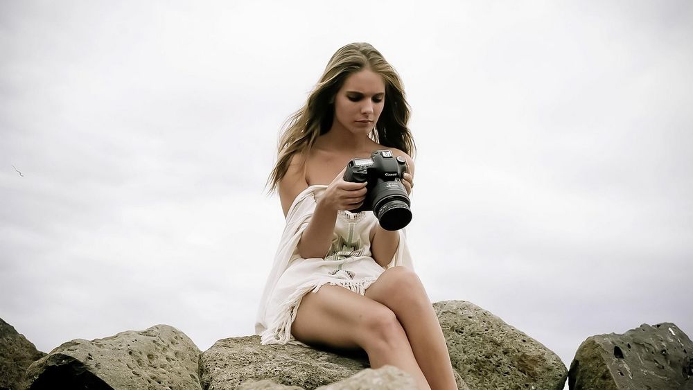 Обои для рабочего стола Полуодетая девушка в белом сидит на камнях с фотоаппаратом на размытом фоне