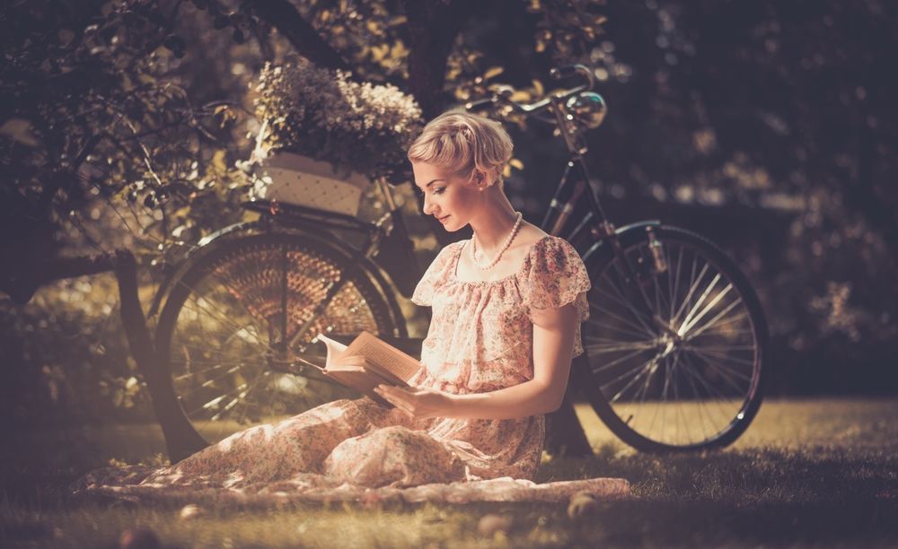 Обои для рабочего стола Девушка сидит на лужайке возле велосипеда и читает книгу на темном размытом фоне