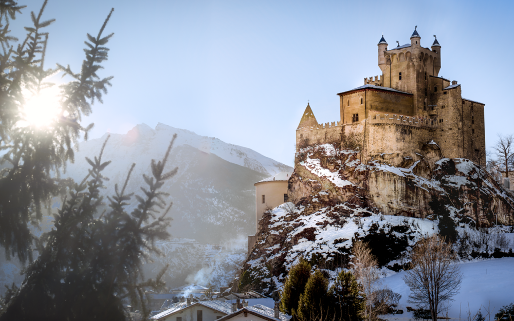 Обои для рабочего стола Заснеженный старинный Castello di Saint-Pierre, Italy / замок Сен-Пьер, Италия на скале на фоне пробивающегося солнца и размытых очертаний гор