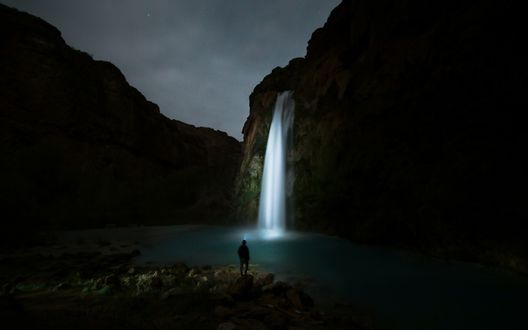 Обои на рабочий стол Мужчина стоит ночью перед водопадом, подсвеченным луной