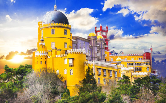 Обои на рабочий стол Дворец Pena Palace, Португалия стоит на скале в лучах заката на фоне ярко-голубого неба с белыми тучками