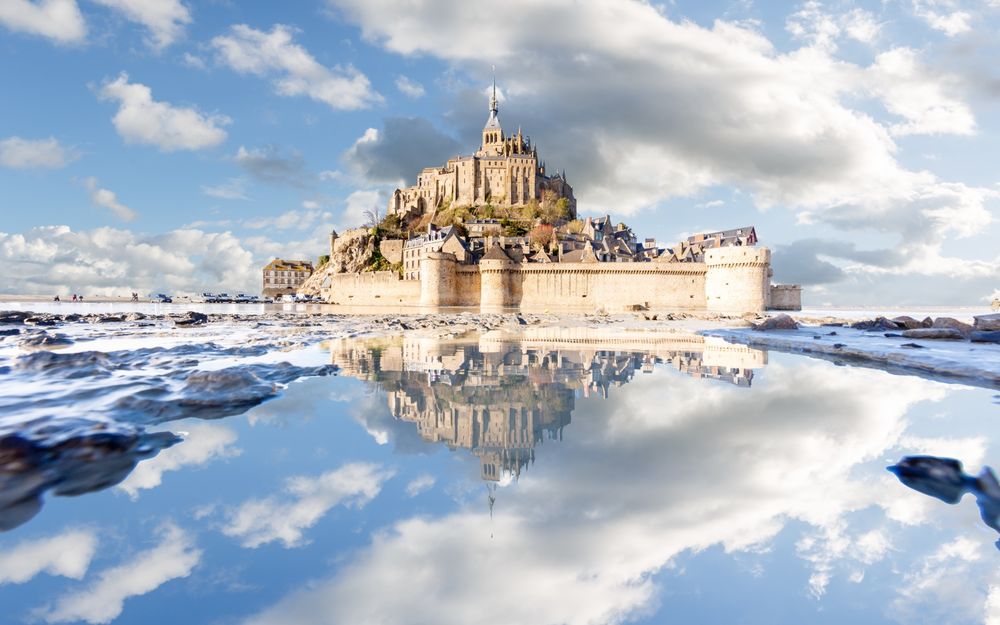 Обои для рабочего стола Остров-крепость Mont Saint-Michel, France / Мон-Сен-Мишель, Франция на фоне ясного голубого неба, белых тучек и яркого солнца, отражается в воде