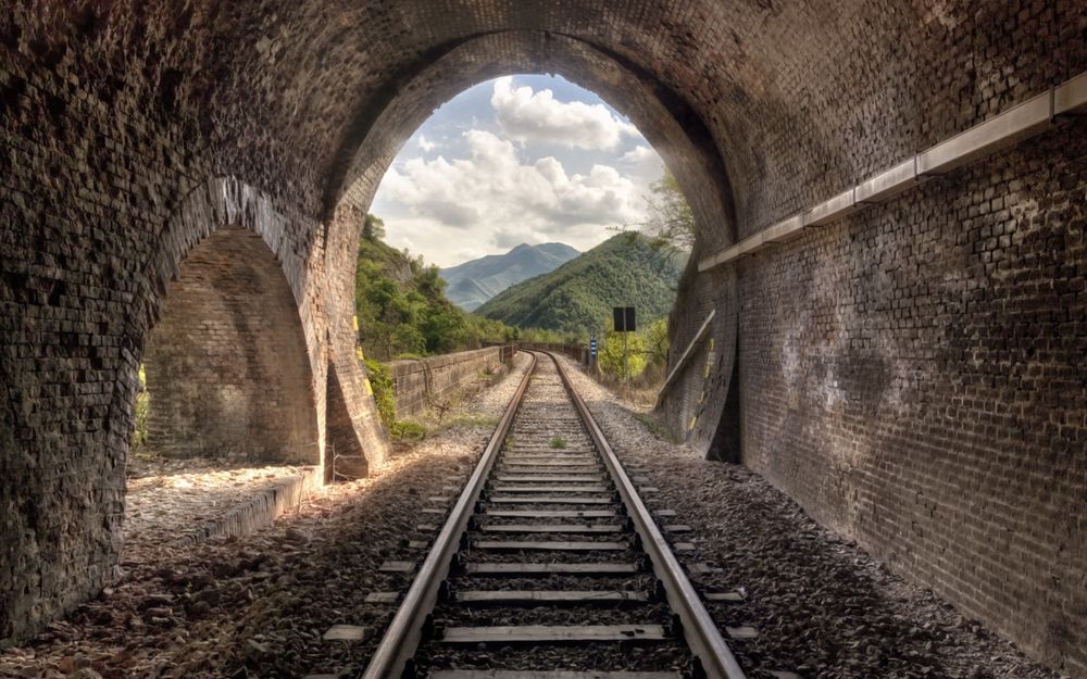 Обои для рабочего стола Железнодорожный туннель в горном массиве, из него виднеются горы на фоне белых туч