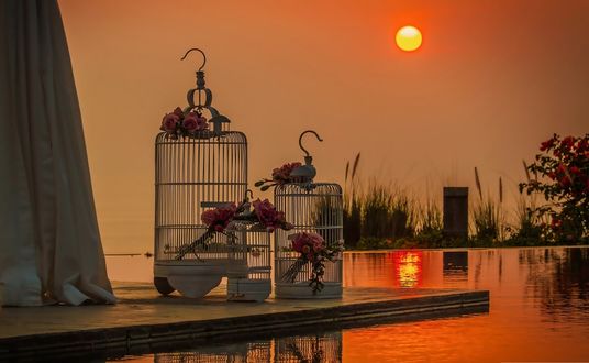 Обои на рабочий стол Пустые клетки с цветами на столике на фоне водоема и заходящего солнца