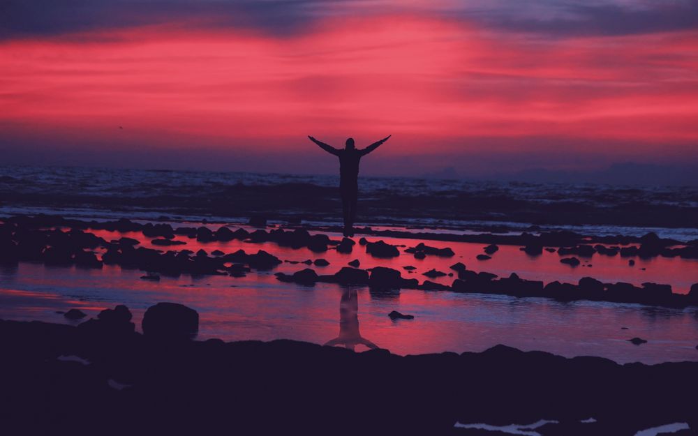 Обои для рабочего стола Силуэт мужчины с раскинутыми руками, стоящего на берегу моря на фоне красного закатного неба