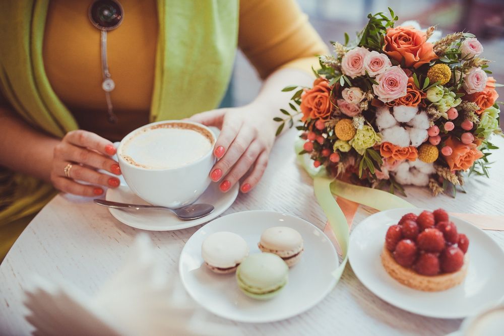 Обои для рабочего стола Девушка сидит за столом и держит в руках чашку капучино, рядом стоит тарелка с печеньем и лежит букет цветов