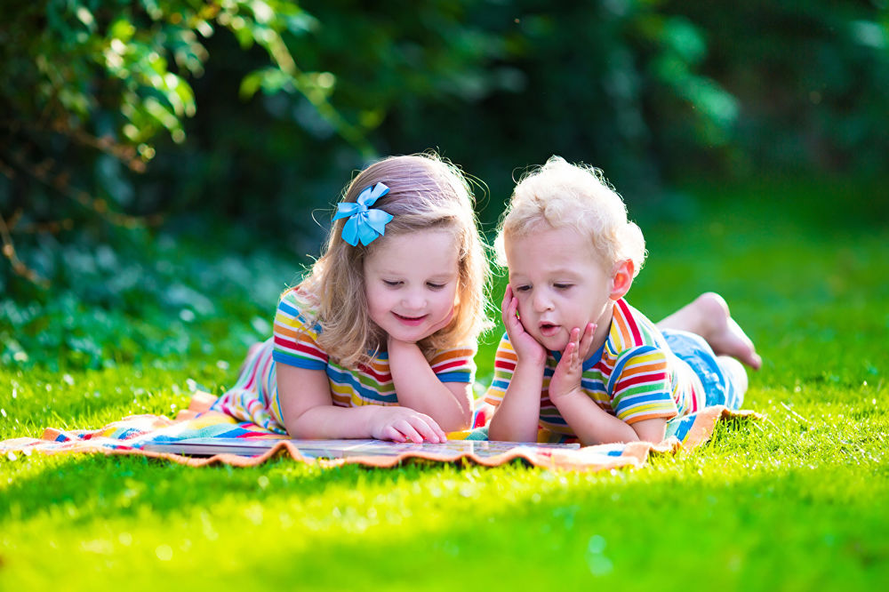 Обои для рабочего стола Дети увлеченно разглядывают картинки в книжке, лежа на траве, в тени под деревьями