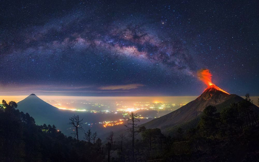 Обои для рабочего стола Извержение вулкана в горах Гватемалы на фоне ночного неба и Млечного пути, вдали виднеются огни города, фотограф Albert Dros