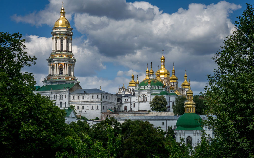 Обои для рабочего стола Комплекс Киево-Печерской Лавры, Киев, Украина виднеется за деревьями на фоне ярко-голубого неба с бело-серыми тучами