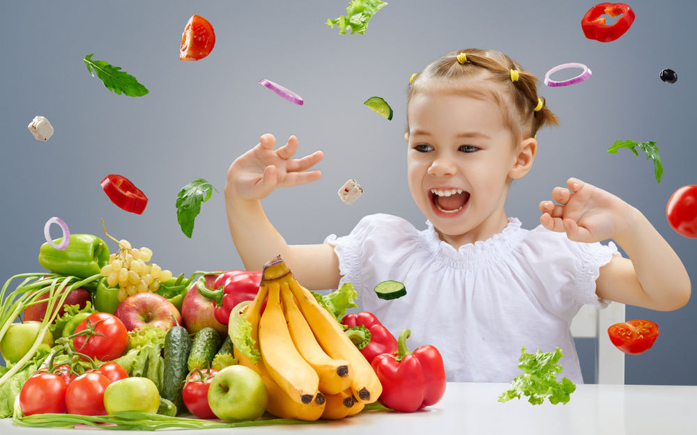 Обои для рабочего стола Девочка сидит за кухонным столом и играется с летающими фруктами и овощами
