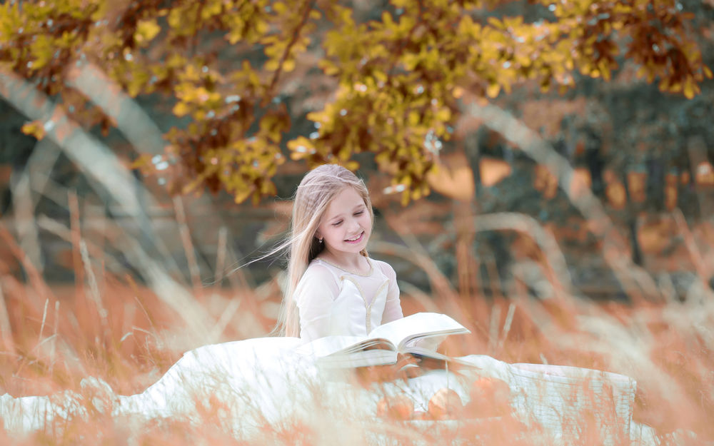 Обои для рабочего стола Светловолосая девочка в белом платье читает книгу, сидя под деревом