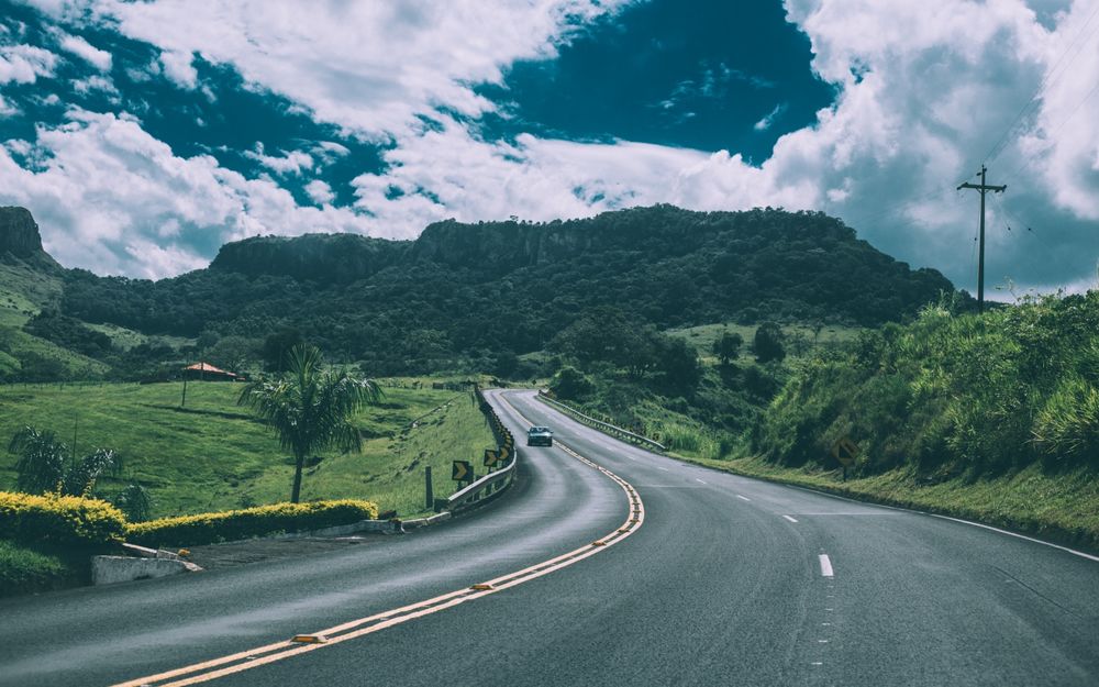 Обои для рабочего стола Машина едет по извилистой асфальтовой дороге по направлению к холмам в пасмурную погоду, фотограф Kaique Rocha
