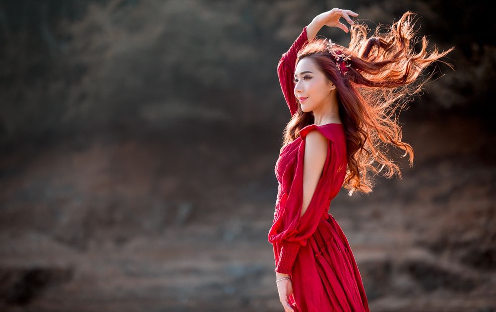 Обои для рабочего стола Азиатка в красном платье с развевающимися волосами стоит на размытом сером фоне