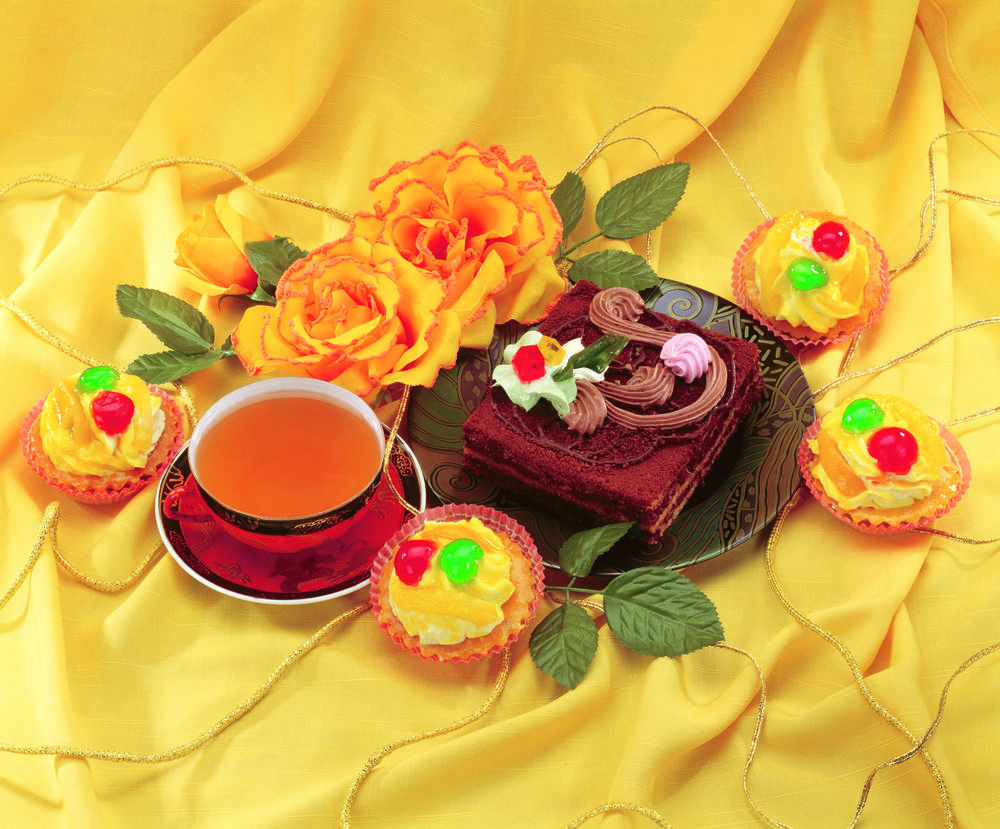 Обои для рабочего стола Шоколадное пирожное на тарелке, маленькие кексы, чашка чая и желтые розы на желтой шелковой ткани