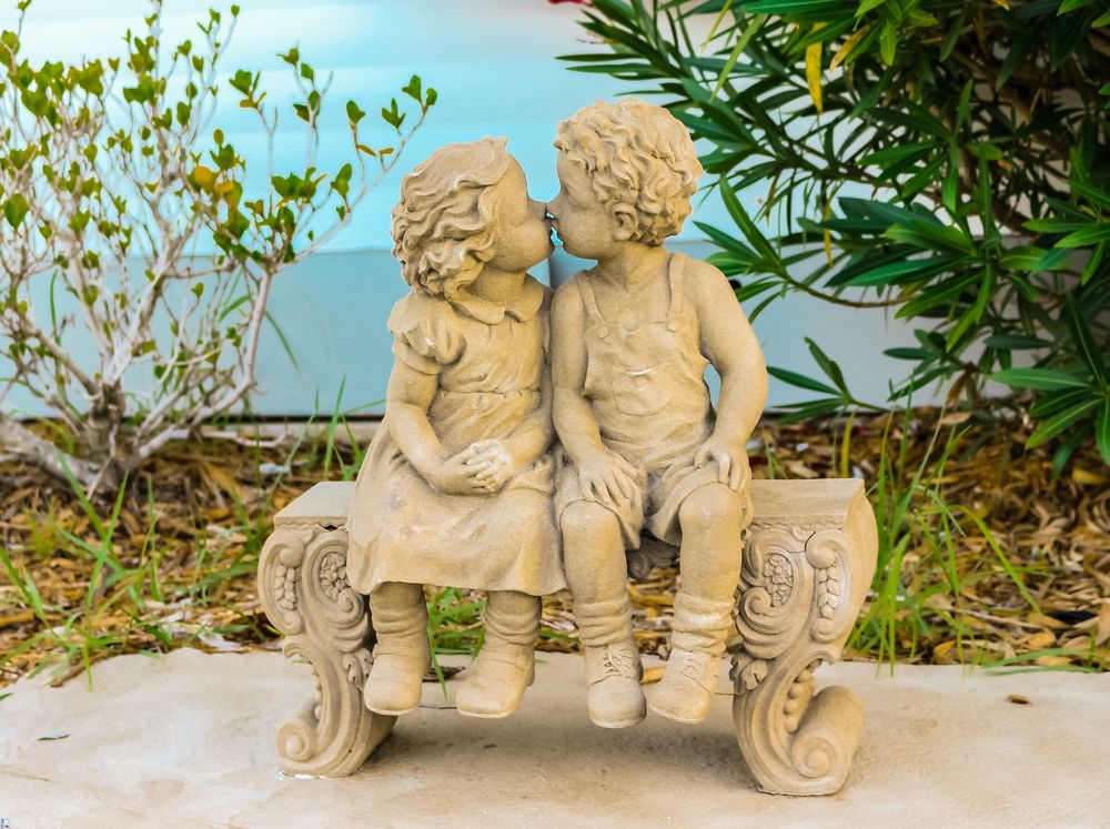 Обои для рабочего стола Скульптуры ангелов целуются на лавочке в парке летом на фоне голубого неба и листьев