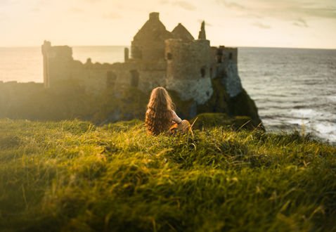 Обои на рабочий стол Девушка сидит в траве на фоне замка Dunluce castle in Northern Ireland / Данлудж в Северной Ирландии, фотограф Lizzy Gadd