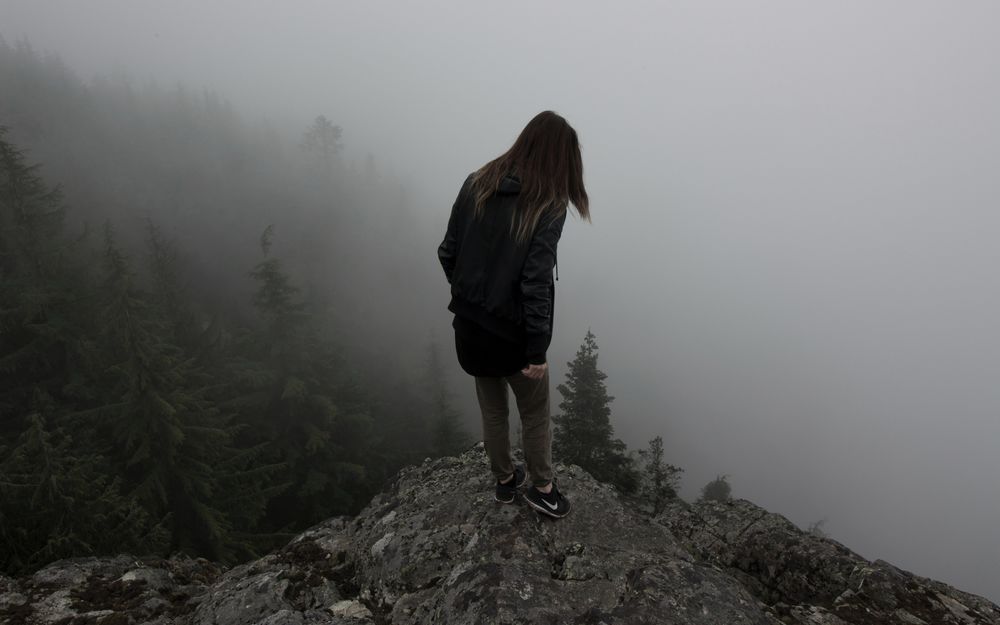 Обои для рабочего стола Девушка в куртке с капюшоном стоит на выступе горы, а вокруг клубится туман