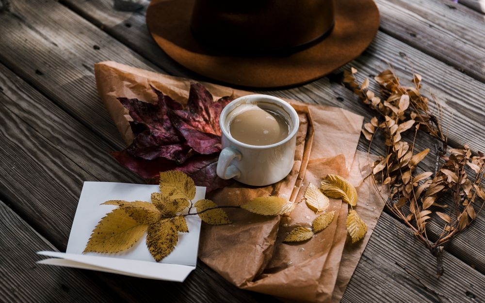 Обои для рабочего стола Чашка кофе стоит на деревянном столе, рядом пожелтевшие осенние листья и коричневая шляпа