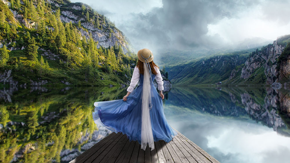 Обои для рабочего стола Девушка в длинном платье в шляпе стоит на мостике на фоне озера у гор