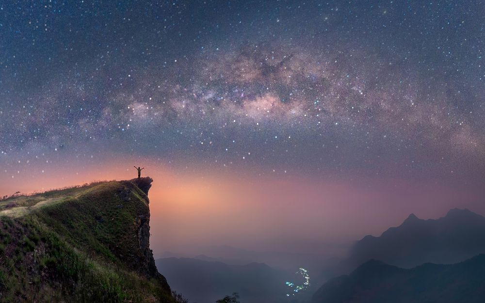 Обои для рабочего стола Человек, раскинув руки, стоит на скале, над туманной долиной, на фоне зарева в звездном небе, by Chaowarin Hadchiang