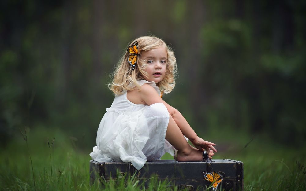 Обои для рабочего стола Девочка в белом платье и с заколкой-бабочкой на волосах сидит на чемодане, фотограф Kim Pennington