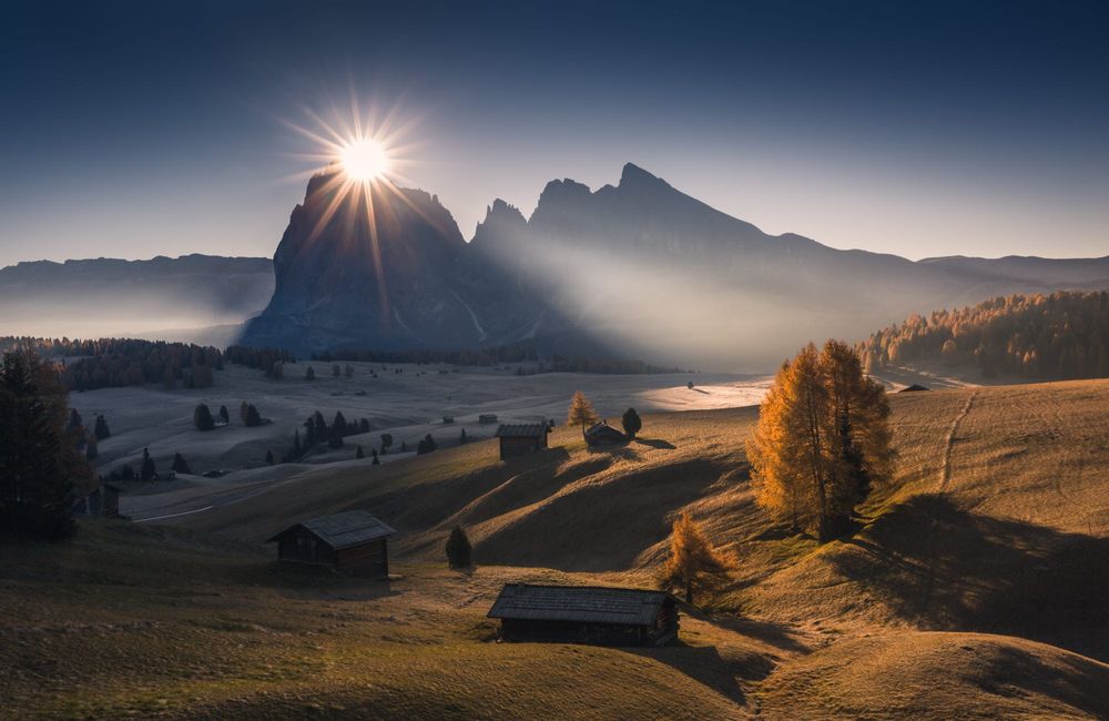 Обои для рабочего стола Солнце над вершиной горы освещает долину с домиками, фотограф Marco Grassi