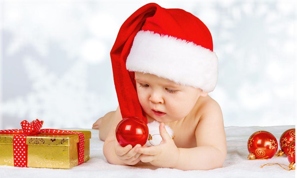 Обои для рабочего стола Младенец в новогодней шапке лежит и играет с новогодними красными шариками на фоне боке