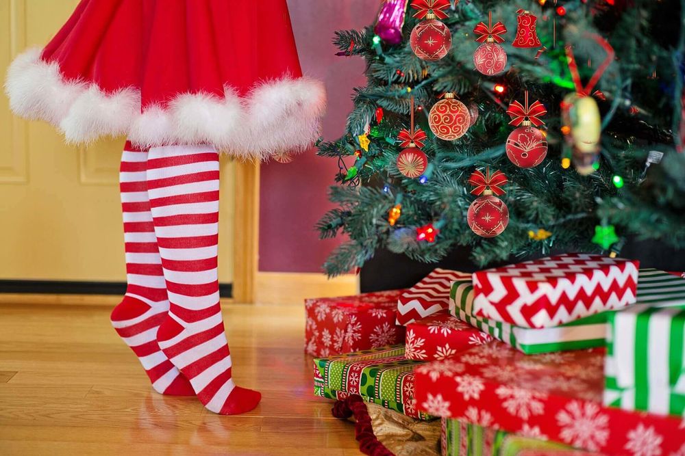 Обои для рабочего стола Девочка в новогоднем наряде стоит на цыпочках и украшает елку, рядом с которой лежат подарки