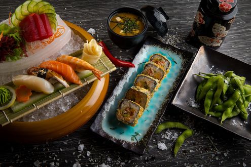 Обои на рабочий стол Японская кухня: суши-ассорти, васаби и имбирь на циновке, нарезанные овощи в миске со льдом, рядом роллы и бобы в черных тарелках