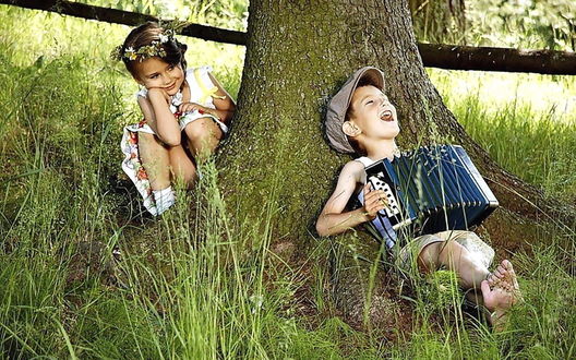 Обои на рабочий стол Мальчик играет на гармошке лежа под деревом, а девочка наблюдает за ним