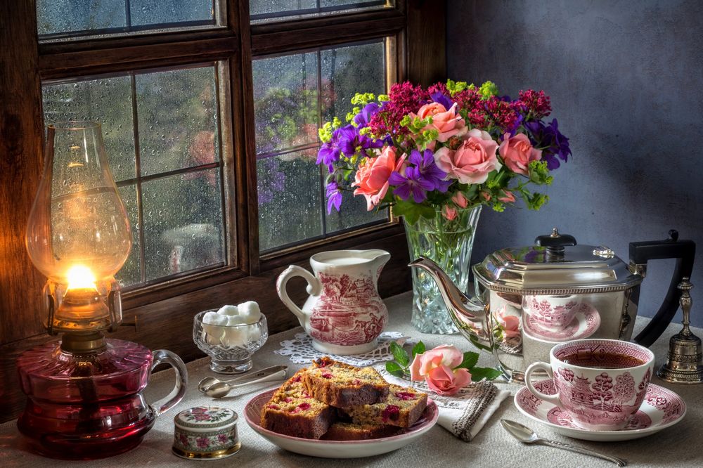 Обои для рабочего стола Стол накрыт для чаепития со свежеиспеченным кексом и стоит букет роз в вазе, все освещено керосиновой лампой, за окном моросит дождь