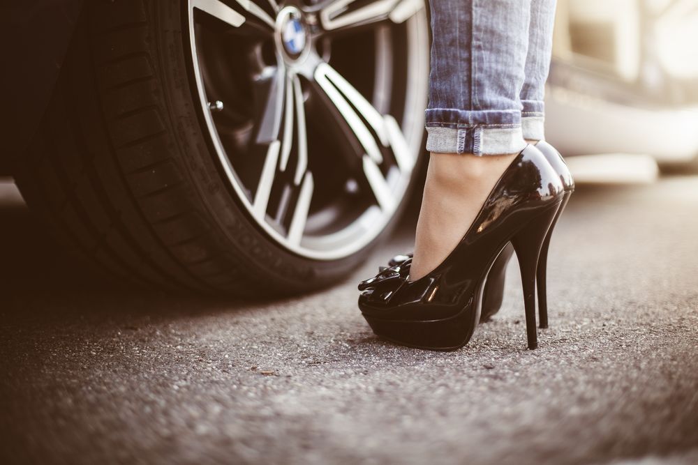 Обои для рабочего стола Женские ножки в черных туфлях стоят около колеса машины