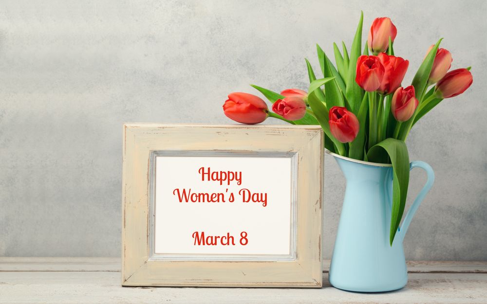 Обои для рабочего стола Букет красных тюльпанов в голубой вазе стоит рядом с рамкой, в которой написано 8 march, Happy Womans day / 8 марта, Международный женский день