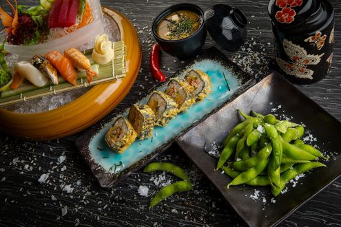 Обои на рабочий стол Японская кухня: суши-ассорти, васаби и имбирь на циновке, нарезанные овощи в миске со льдом, рядом роллы и бобы в черной тарелке